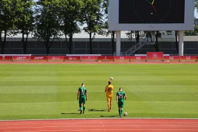 Foto: Trīs futbolisti  dodās ar futbola bumbām un spēles laukumu, zaļš mauriņš