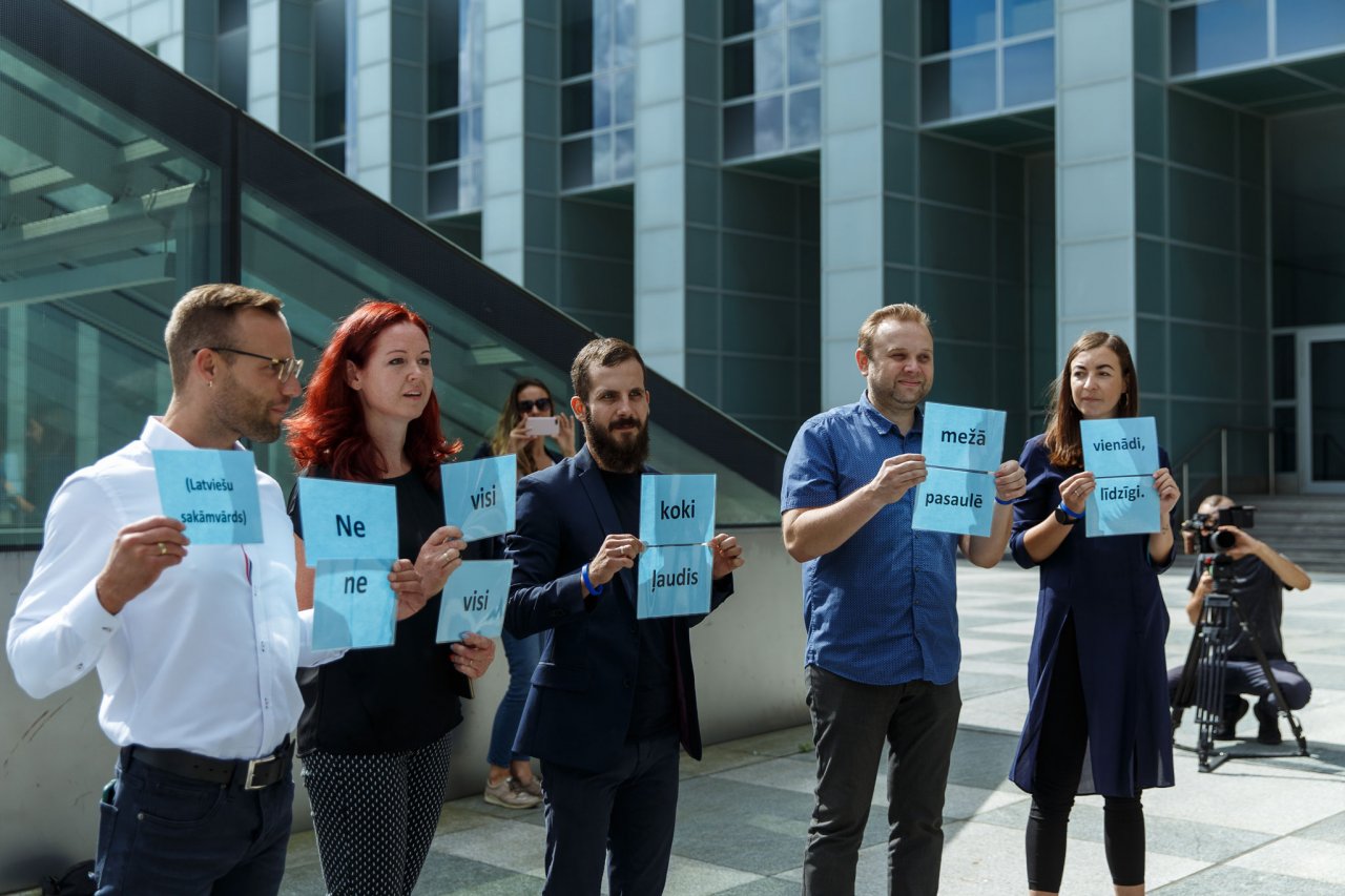 Pieci cilvēki vasarā, Latvijas Nacionālās bibliotēkas fonā, stāv un rokās tur lapas ar tekstu