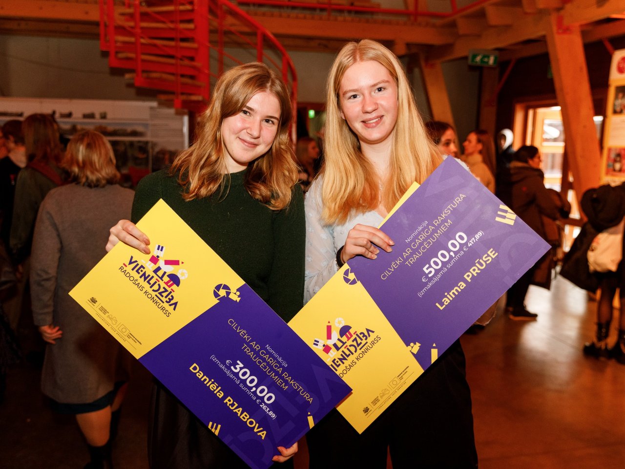 Divas skolnieces smaida un demonstrē savas balvas - čekus par uzvaru konkursā